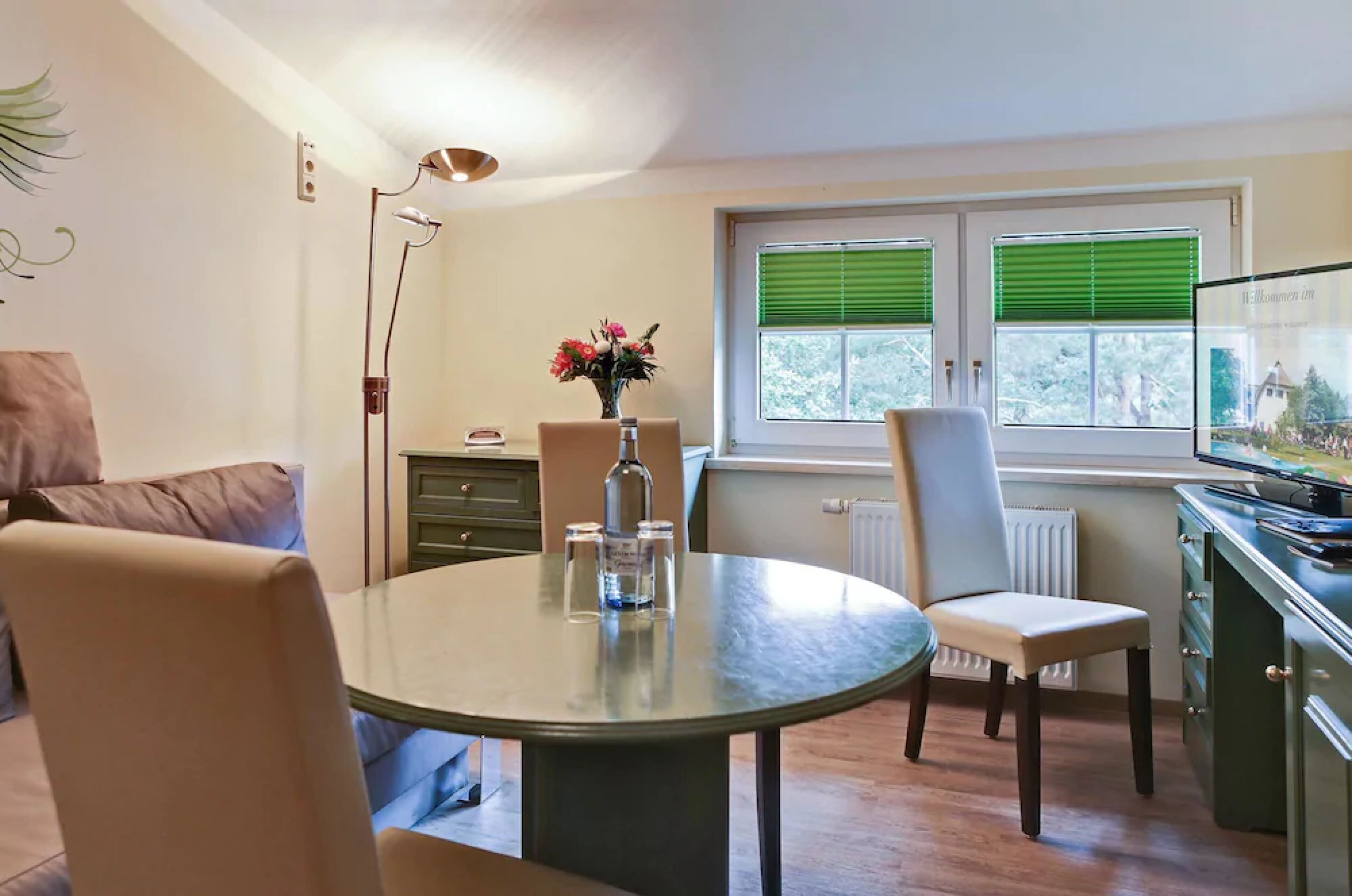 A dining area with a round table, chairs, a vase with flowers, and a window with blinds.
