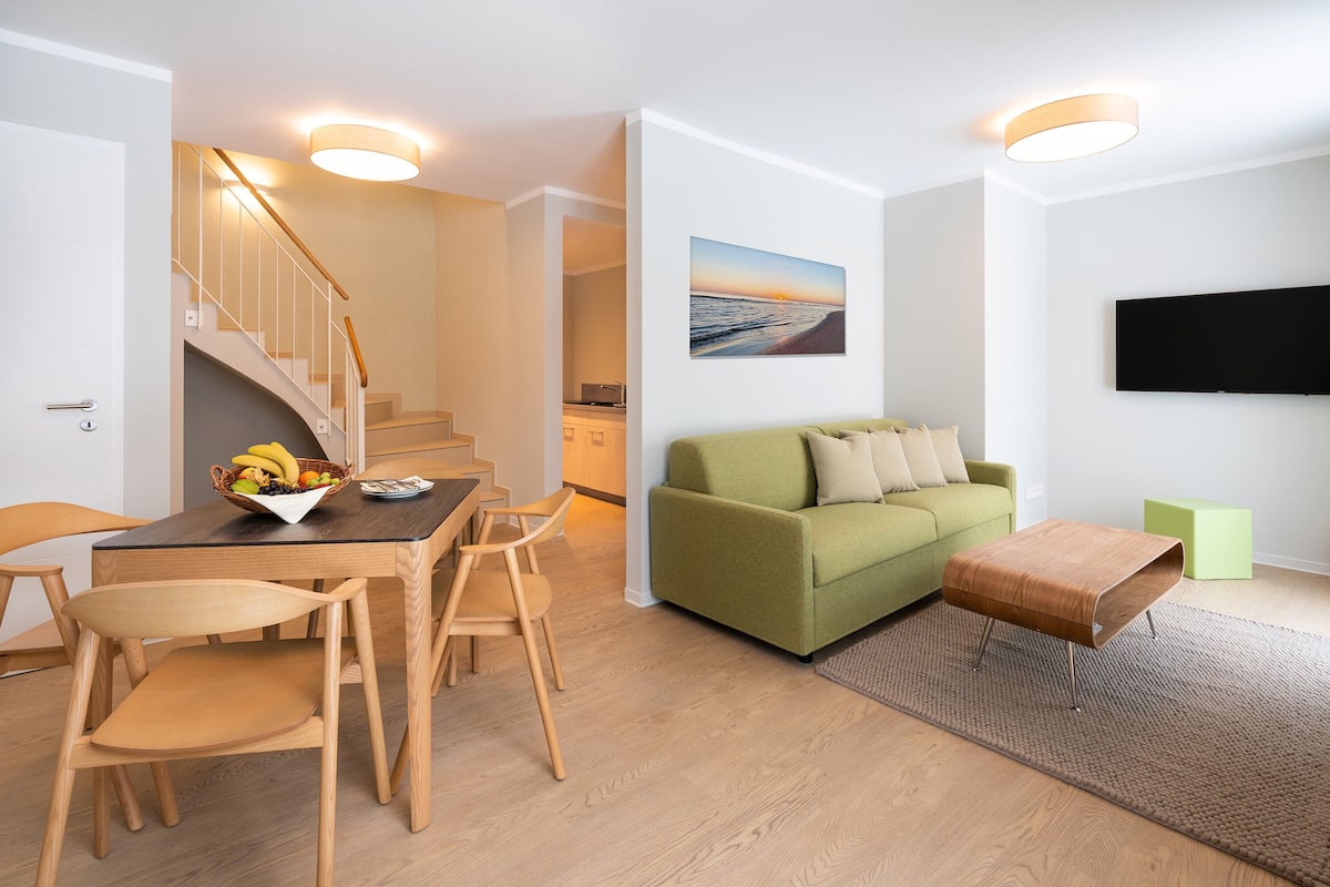 Uma sala de estar moderna com um sofá verde, mesa de jantar de madeira e cadeiras. Há uma escada que leva a outro cômodo, uma TV de tela plana na parede e um quadro sobre o sofá.