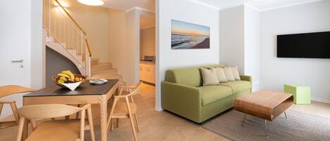 Ein modernes Wohnzimmer mit einem grünen Sofa, einem Holzesstisch und Stühlen. Eine Treppe führt in einen anderen Raum, ein Flachbildfernseher ist an der Wand montiert und über dem Sofa hängt ein Gemälde.