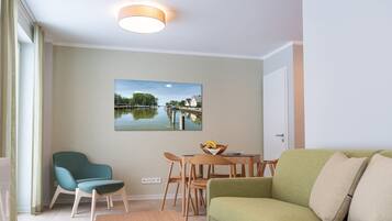 A modern living room with a green sofa, teal armchair, and wooden dining table with chairs. A painting of a water scene is on the wall.