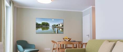 Una sala moderna con un sofá verde, un sillón azul verdoso y una mesa de comedor de madera con sillas. En la pared, un cuadro de una escena acuática.