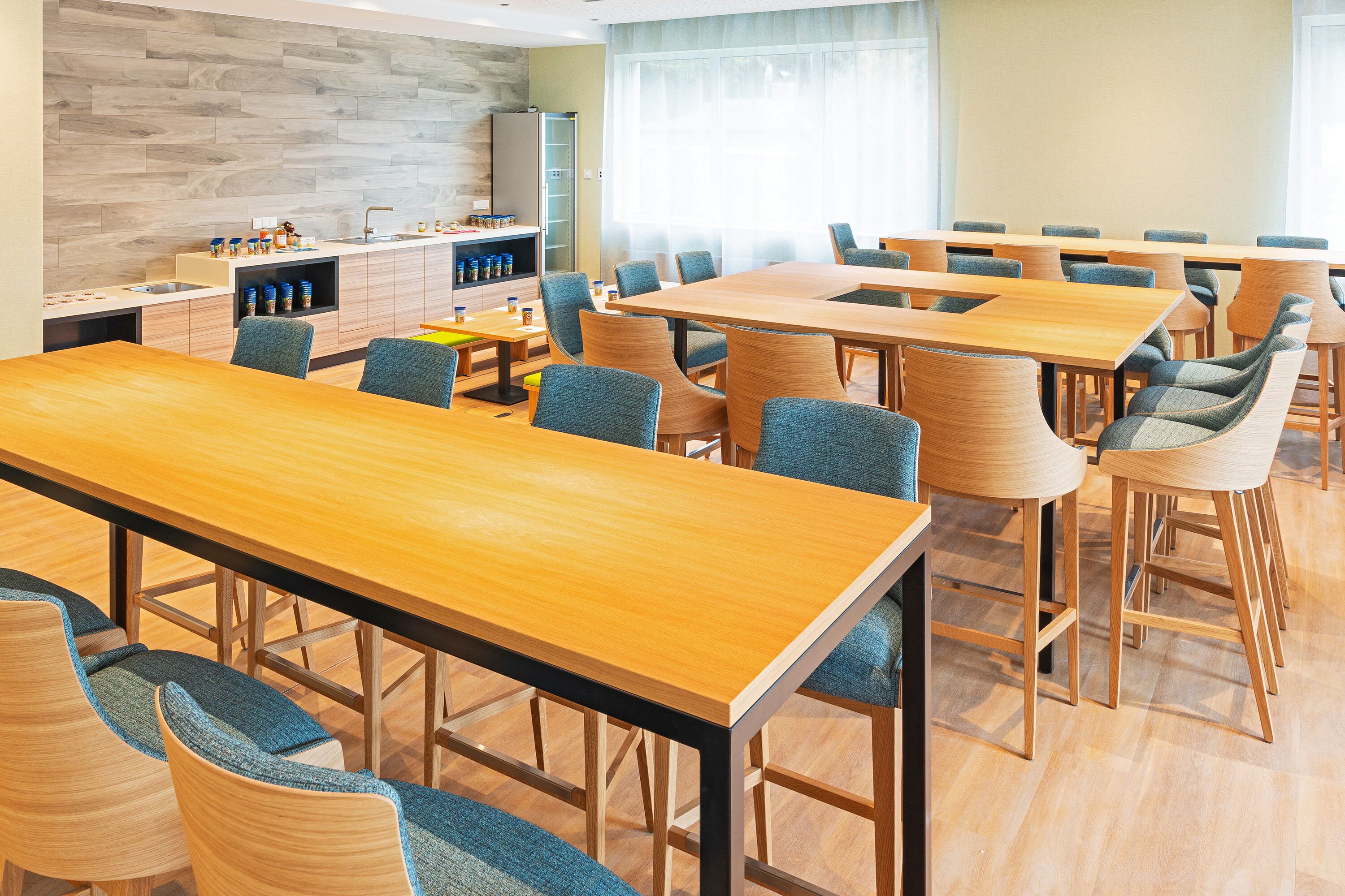 A modern dining area with multiple tables and chairs, a kitchen area with a wooden backsplash, and a large window providing natural light.