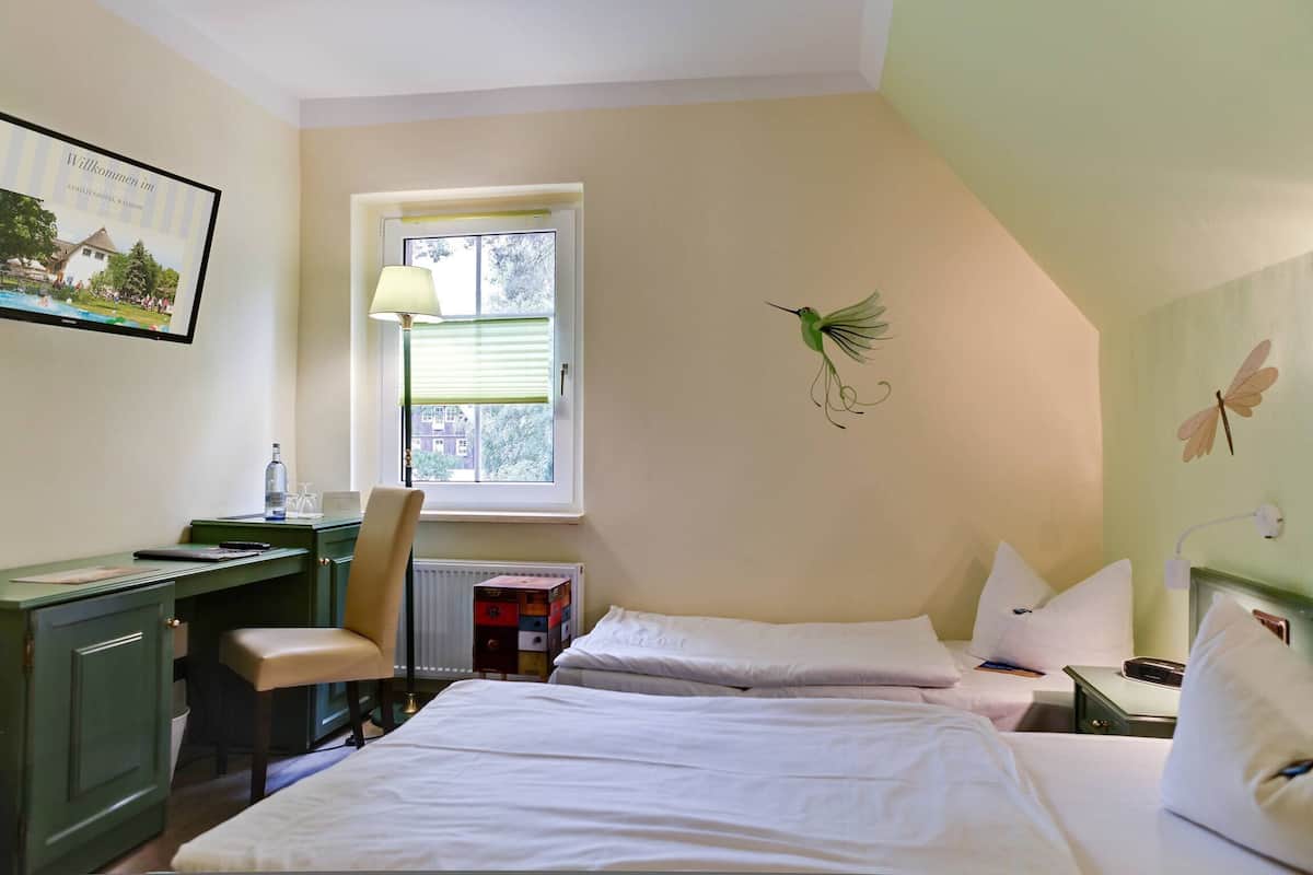 Quarto de hotel com duas camas, uma secretária, uma cadeira, janela com persianas, radiador e luminária na parede.
