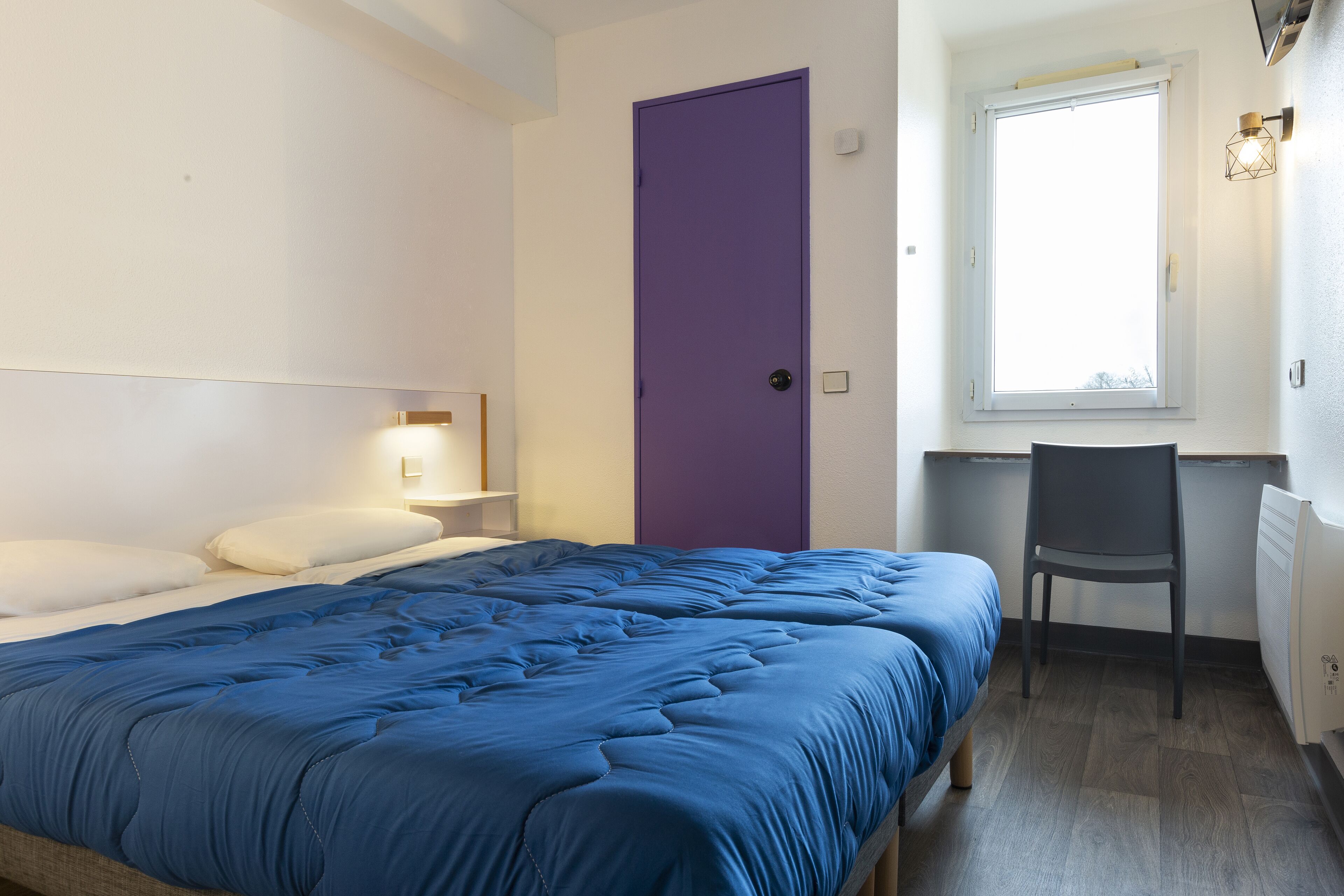 A bedroom with a bed, a desk, a chair, a purple door, and a window.