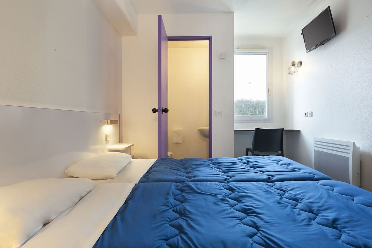 Une chambre à coucher comprenant un lit, une table de chevet, un bureau, une chaise, un radiateur et un téléviseur fixé au mur.
