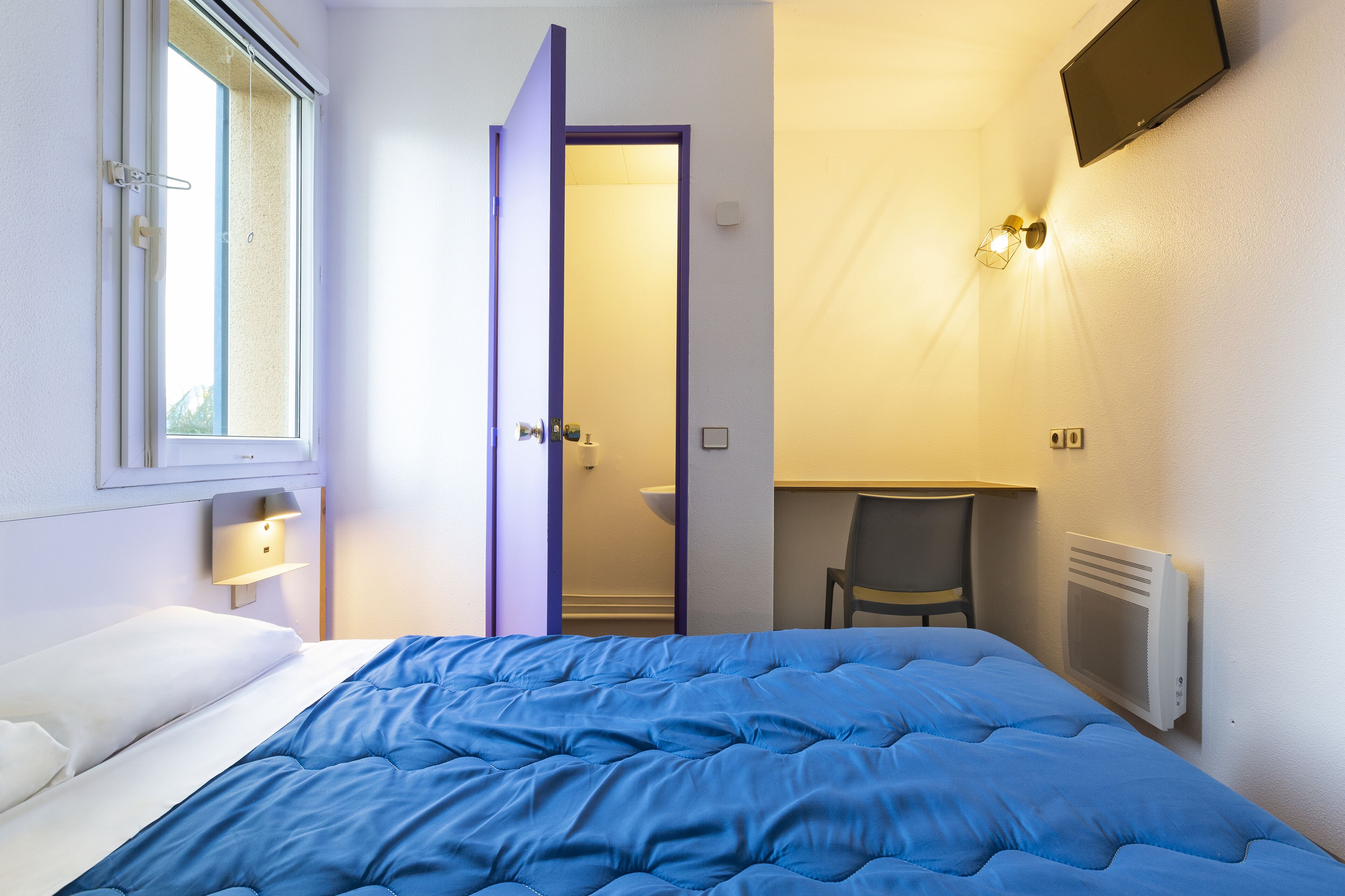 A hotel room with a bed, a desk, a chair, a TV, a window, and a bathroom door.