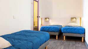 Une chambre avec deux lits simples, chacun doté d’un matelas bleu et d’un cadre en bois. On y trouve des tables de chevet avec des lampes, et la chambre est recouverte d’un parquet.