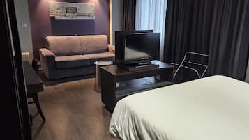 A hotel room with a bed, a desk with a television, a sofa, and a window with curtains.