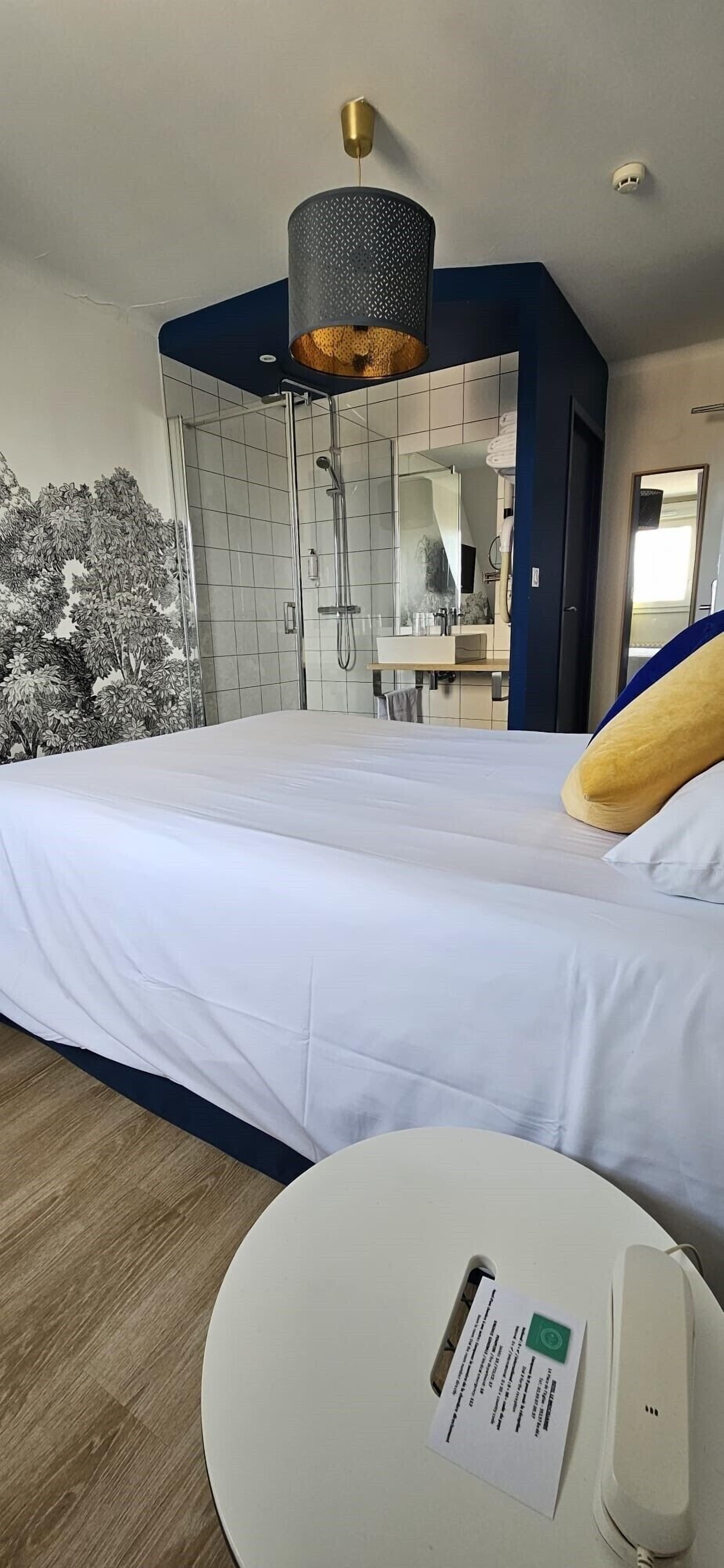 Habitación de hotel con cama, una mesa redonda con teléfono y una tarjeta, zona de ducha y un espejo.