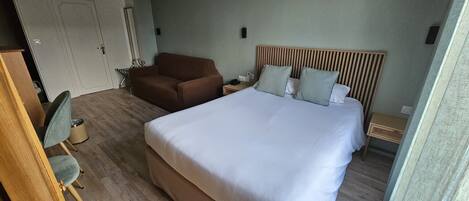 Una habitación de hotel con una cama grande, un sillón marrón, una mesita de noche de madera y una puerta.