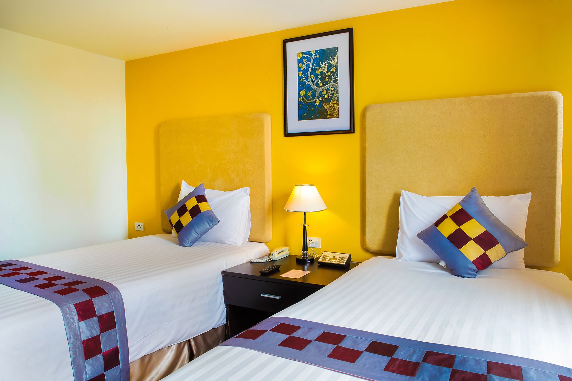 Habitación de hotel con dos camas, una mesita de noche con lámpara, teléfono y un cuadro en la pared.