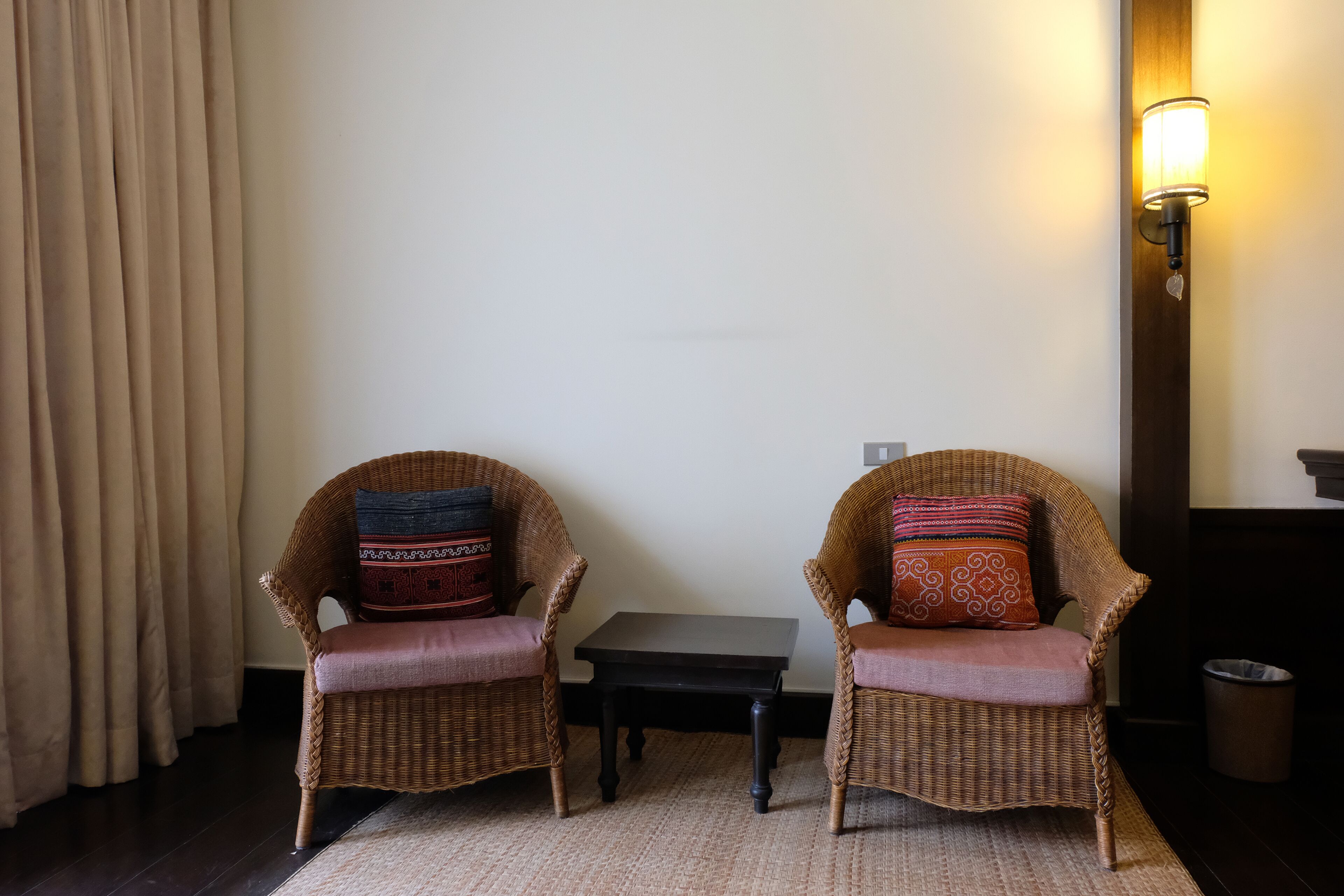Two wicker chairs with cushions, a small table, and a wall-mounted light fixture in a room with a dark floor and light-colored walls.