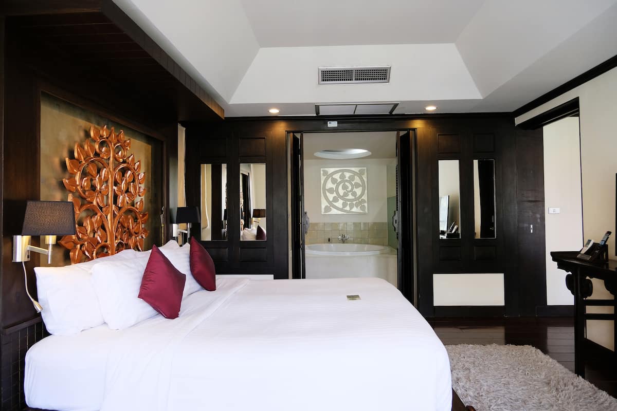 A hotel room with a large bed, a wooden accent wall, a bathroom with a bathtub, and a small desk with a lamp.
