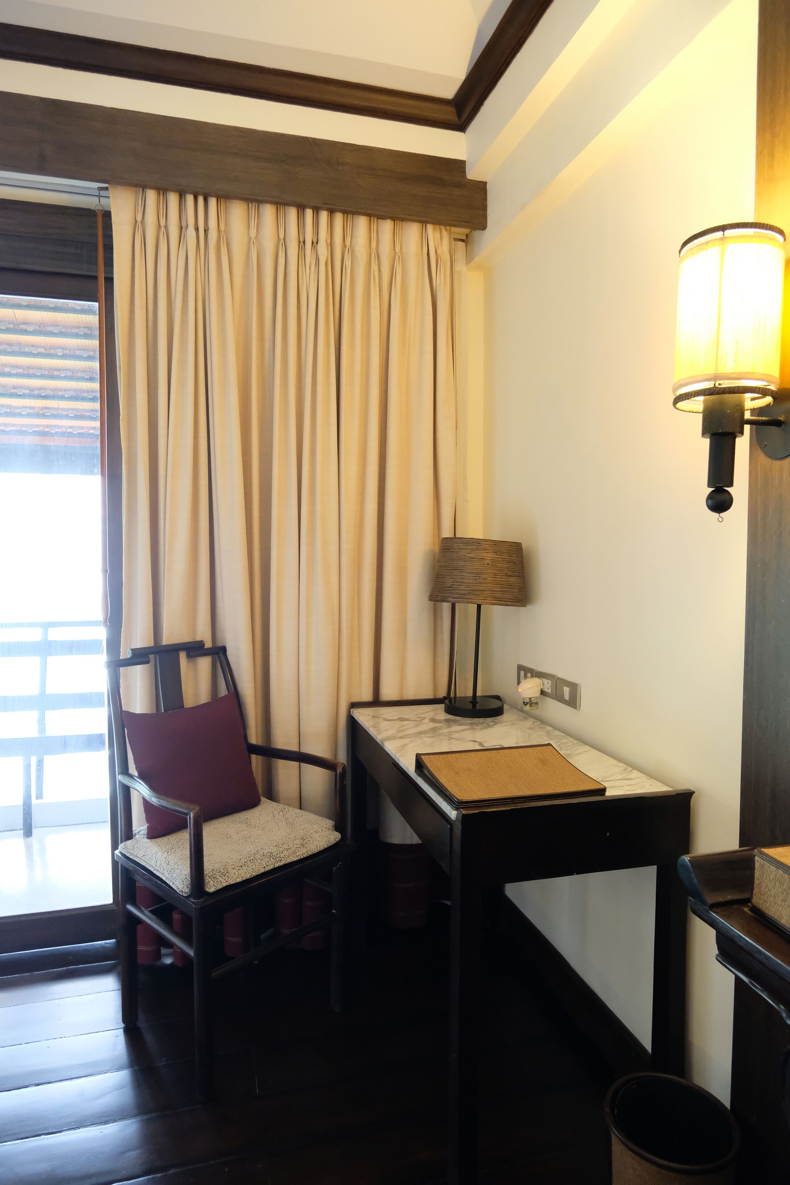 Habitación de hotel con piso de madera, un escritorio con lámpara, una silla con cojín y una ventana con cortinas.