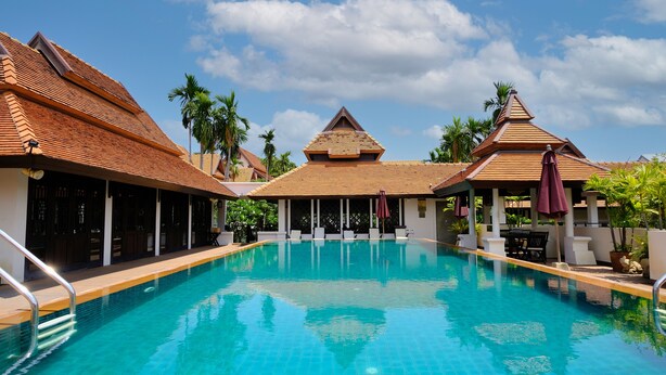 A swimming pool with a tiled deck, surrounded by covered seating areas and palm trees.