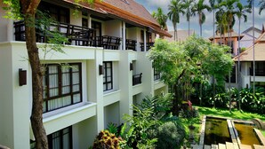 A multi-story hotel with balconies, a garden with palm trees, and a small water feature.