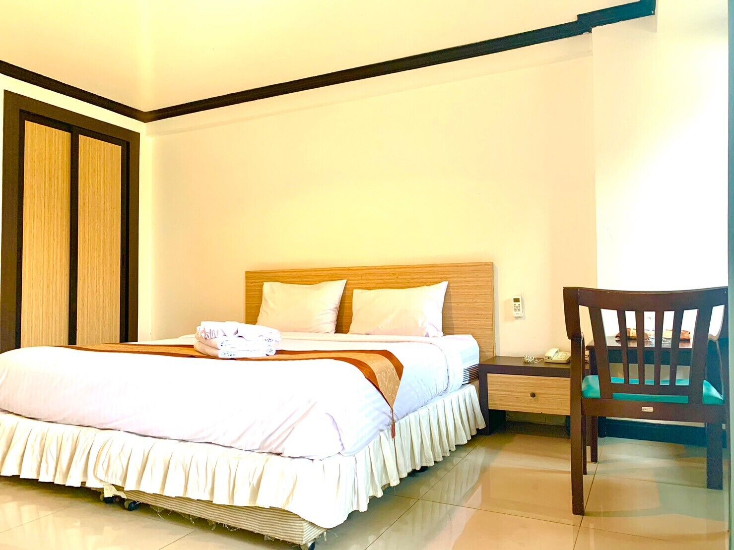 A neatly made bed with a wooden headboard, a bedside table with a lamp, and a chair in a well-lit room with a tiled floor.