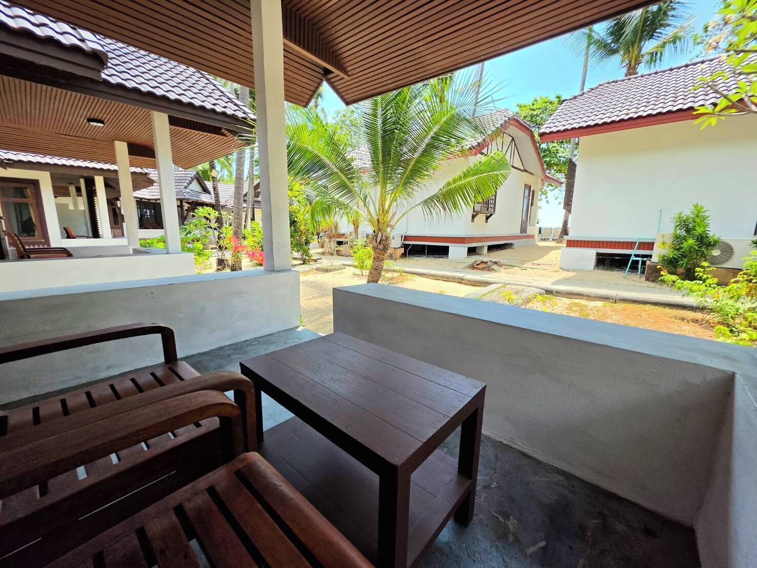 A covered outdoor seating area with a wooden bench and a table, overlooking a courtyard with palm trees and a building with a red roof.