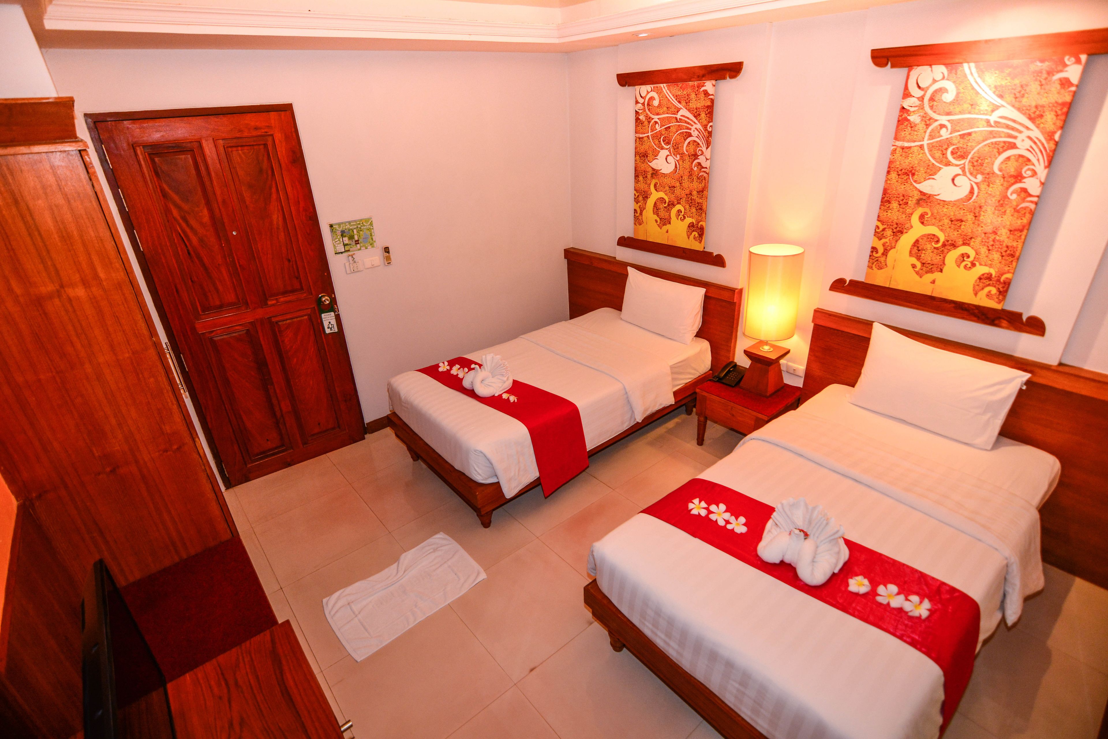 A hotel room with two single beds, a wooden door, a nightstand with a lamp, and a wall with decorative panels.