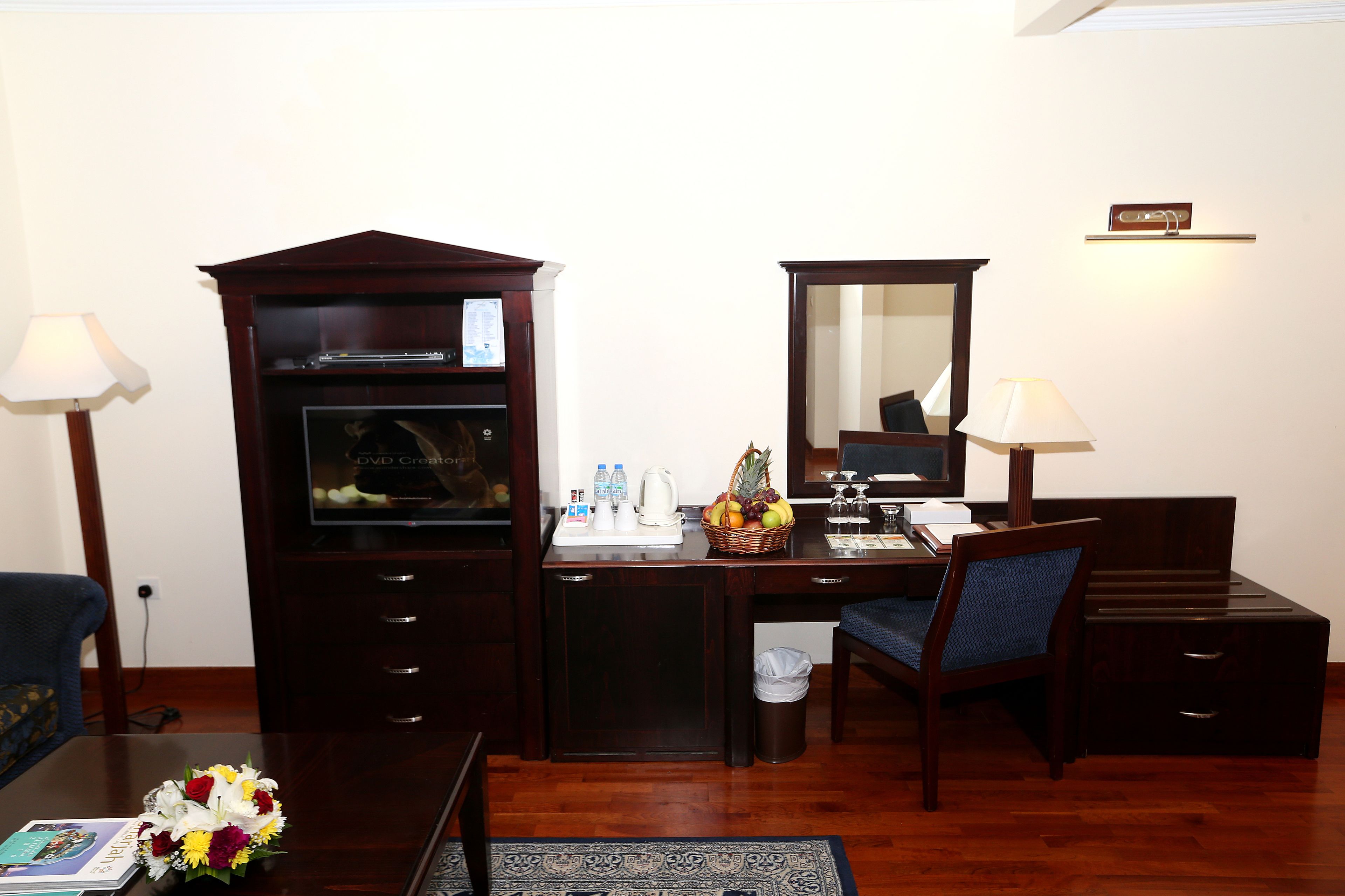 A hotel room with a wooden desk, a chair, a small trash bin, a desk lamp, a mirror, and a television.