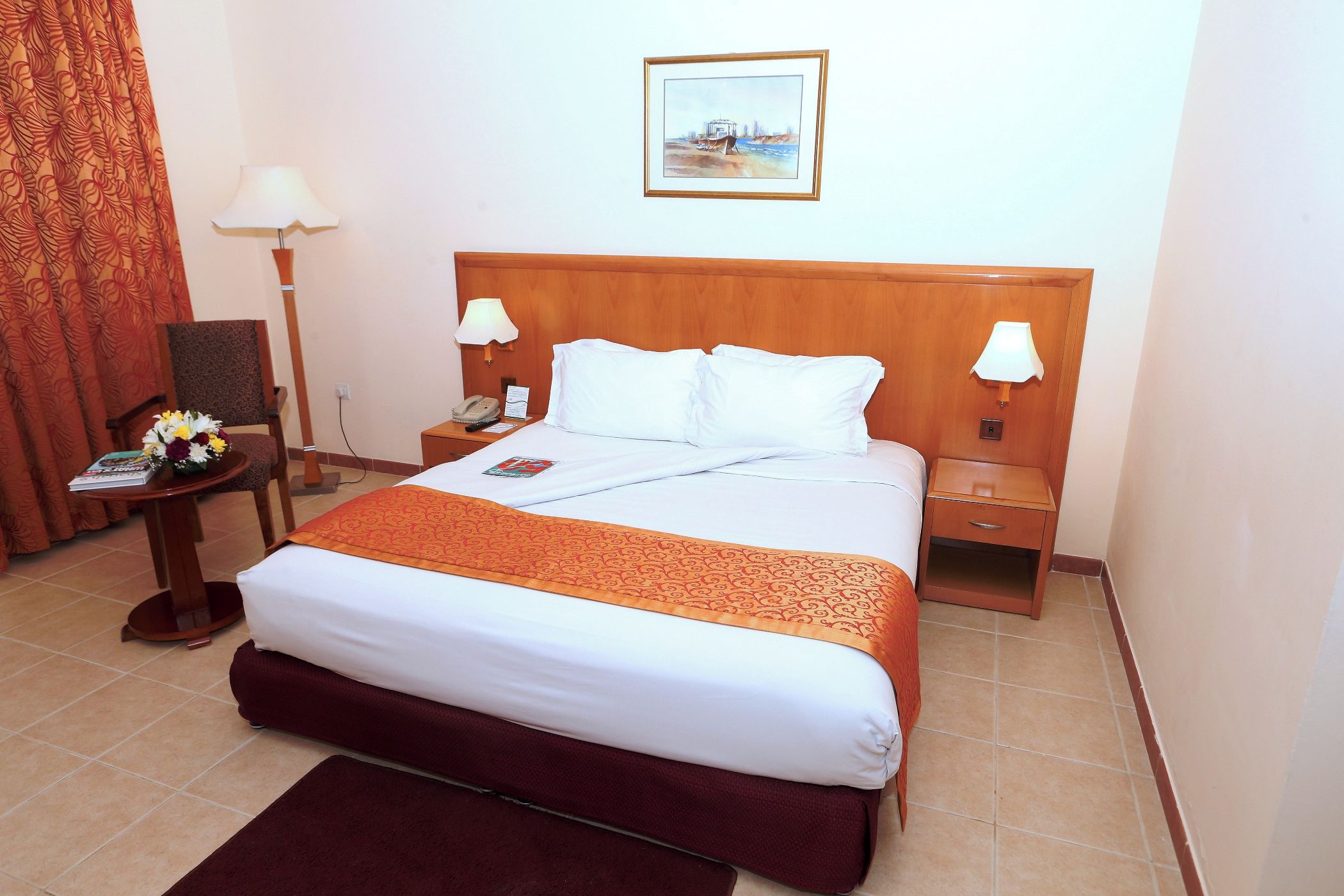 A hotel room with a large bed, two bedside tables, a nightstand, a chair, a lamp, a framed picture on the wall, and a window with curtains.
