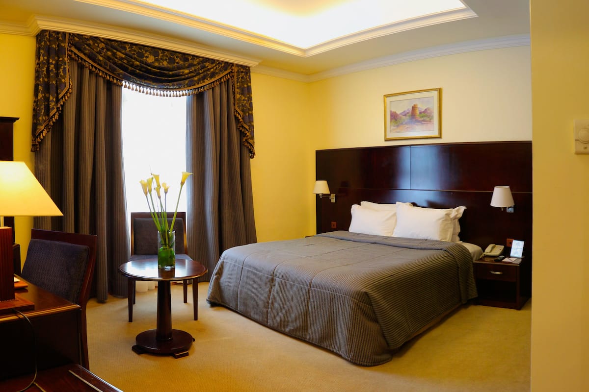 Ein Hotelzimmer mit einem großen Bett, einem Schreibtisch, einem Stuhl, einem kleinen Tisch mit Blumenvase und einem Bild an der Wand.