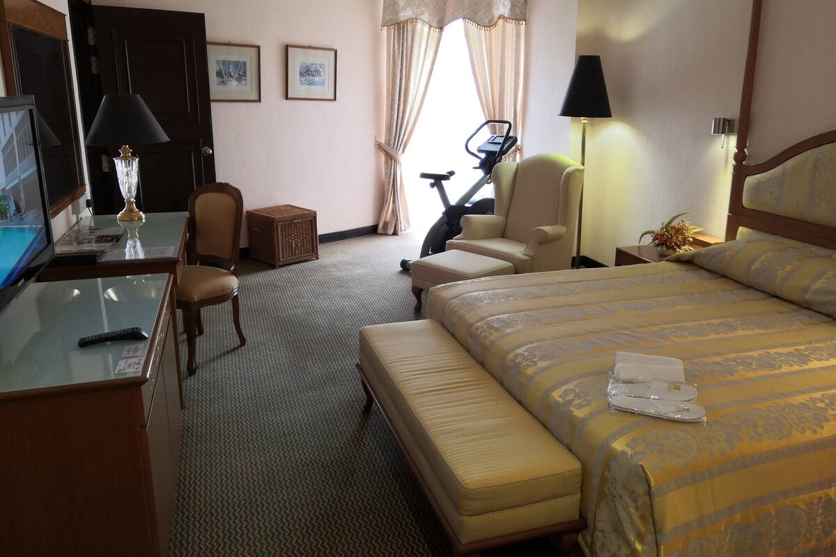 A hotel room with a bed, a desk with a TV, a chair, a small table, a lamp, and a bicycle.