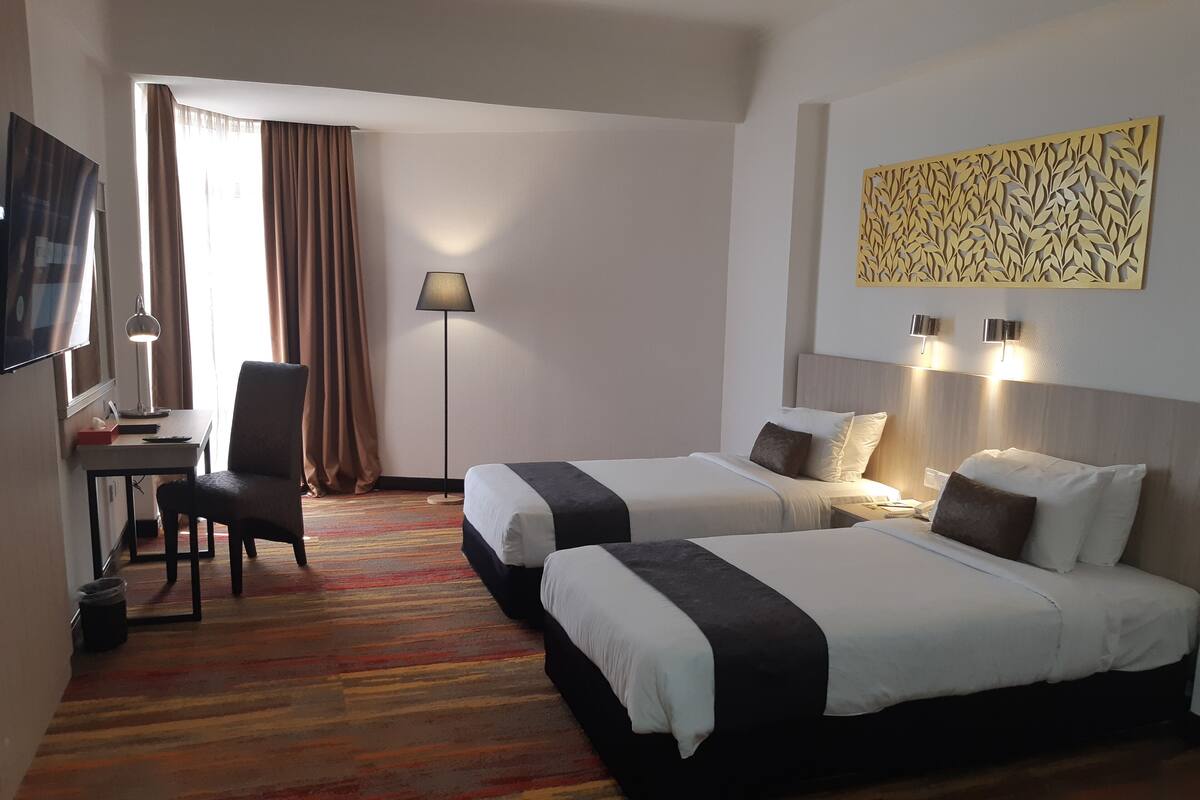 A hotel room with two beds, a desk with a chair, a lamp, and a decorative wall panel.