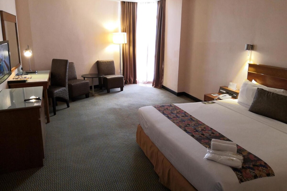 A hotel room with a bed, a desk, a chair, a small table, and a lamp.