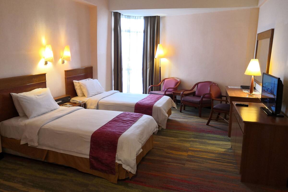 A hotel room with two beds, a desk with a chair, a television, and two lamps.