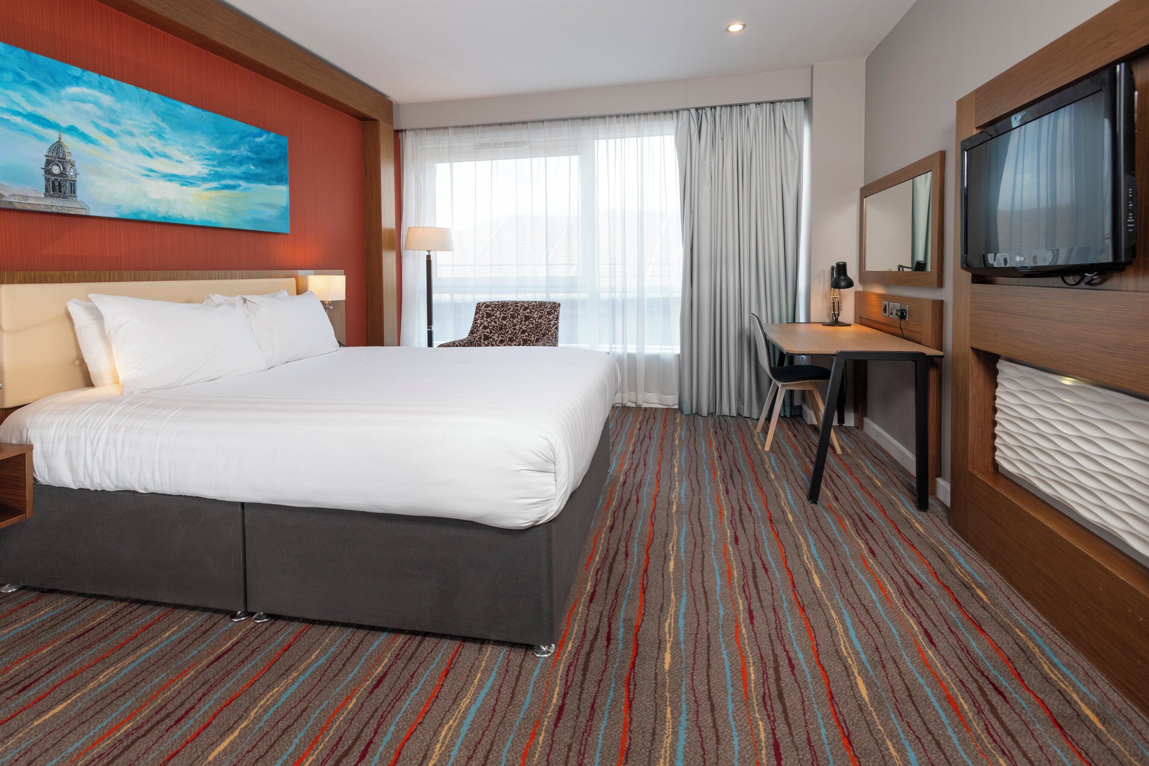 A hotel room with a large bed, a desk, a television, and a window with curtains.