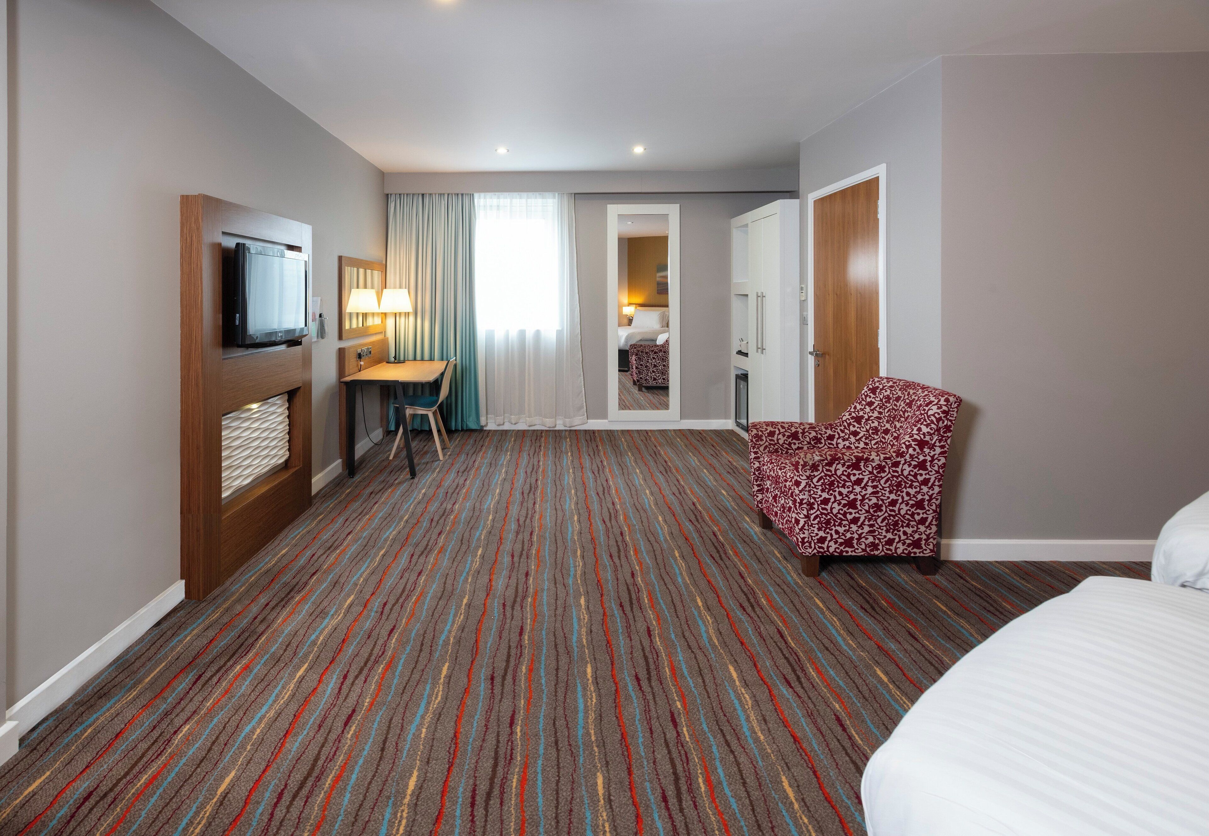 A hotel room with a TV, a desk with a chair, a patterned armchair, and a bed.