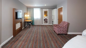 A hotel room with a TV, a desk with a chair, a patterned armchair, and a bed.