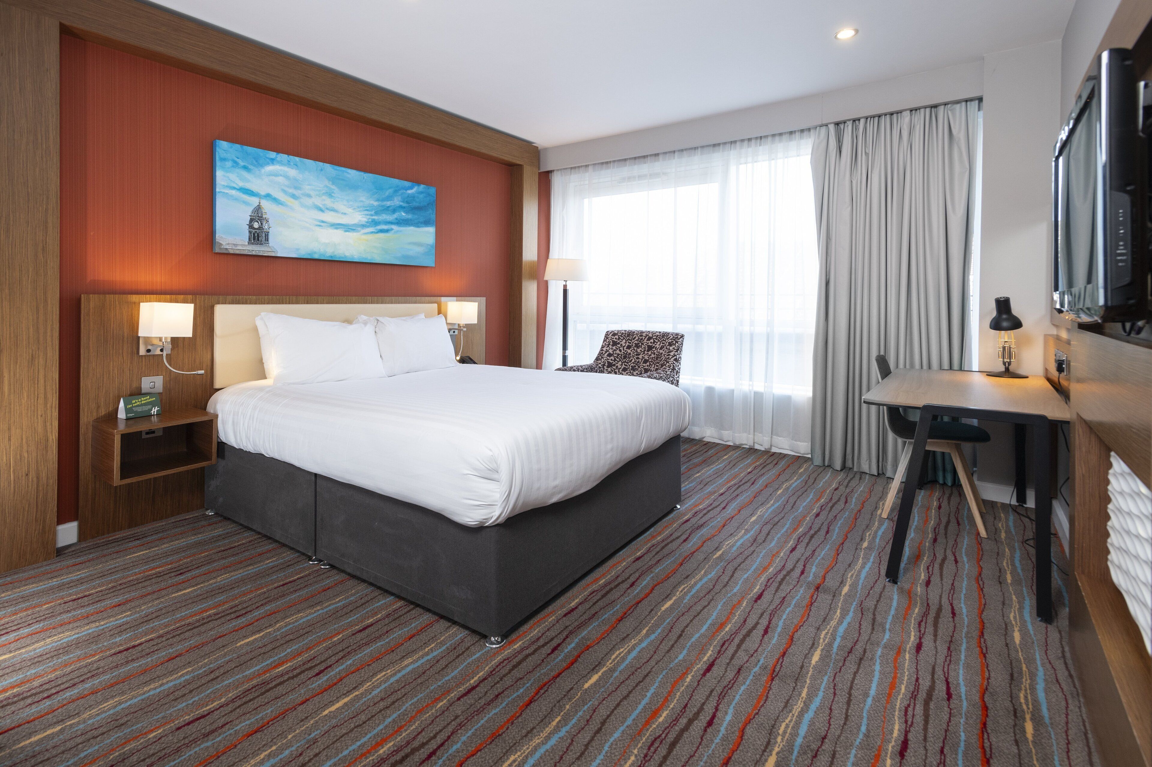 A hotel room with a large bed, a desk, a chair, a TV, and a painting on the wall.