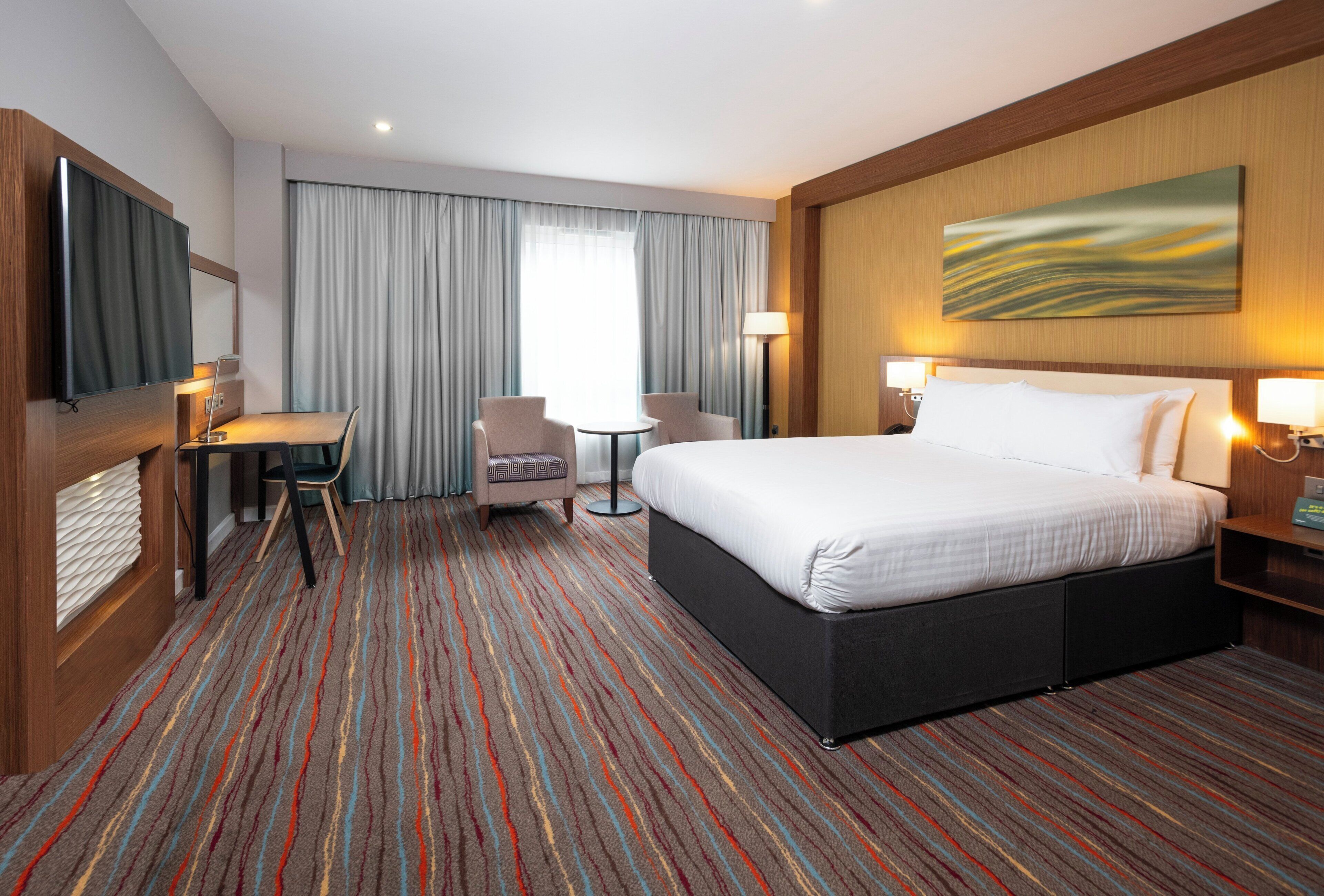 Habitación de hotel con una cama grande, un escritorio, una silla, una mesita y un televisor.