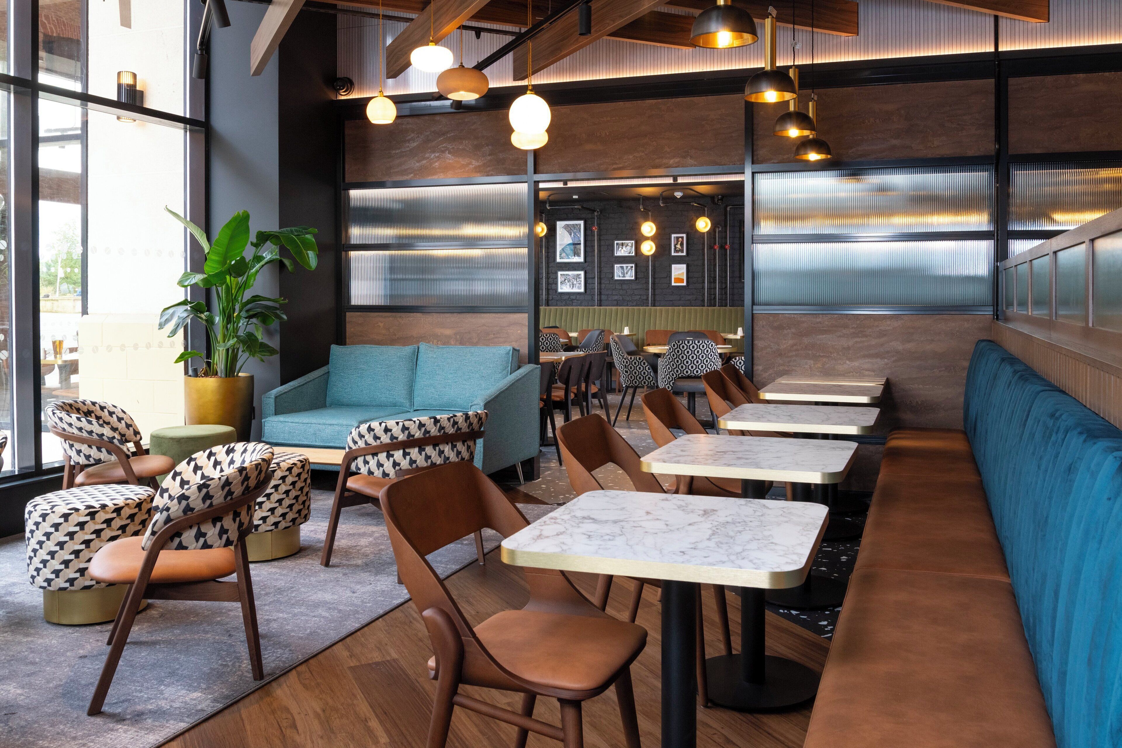 A modern restaurant interior with wooden tables, patterned chairs, a blue sofa, and a large plant.