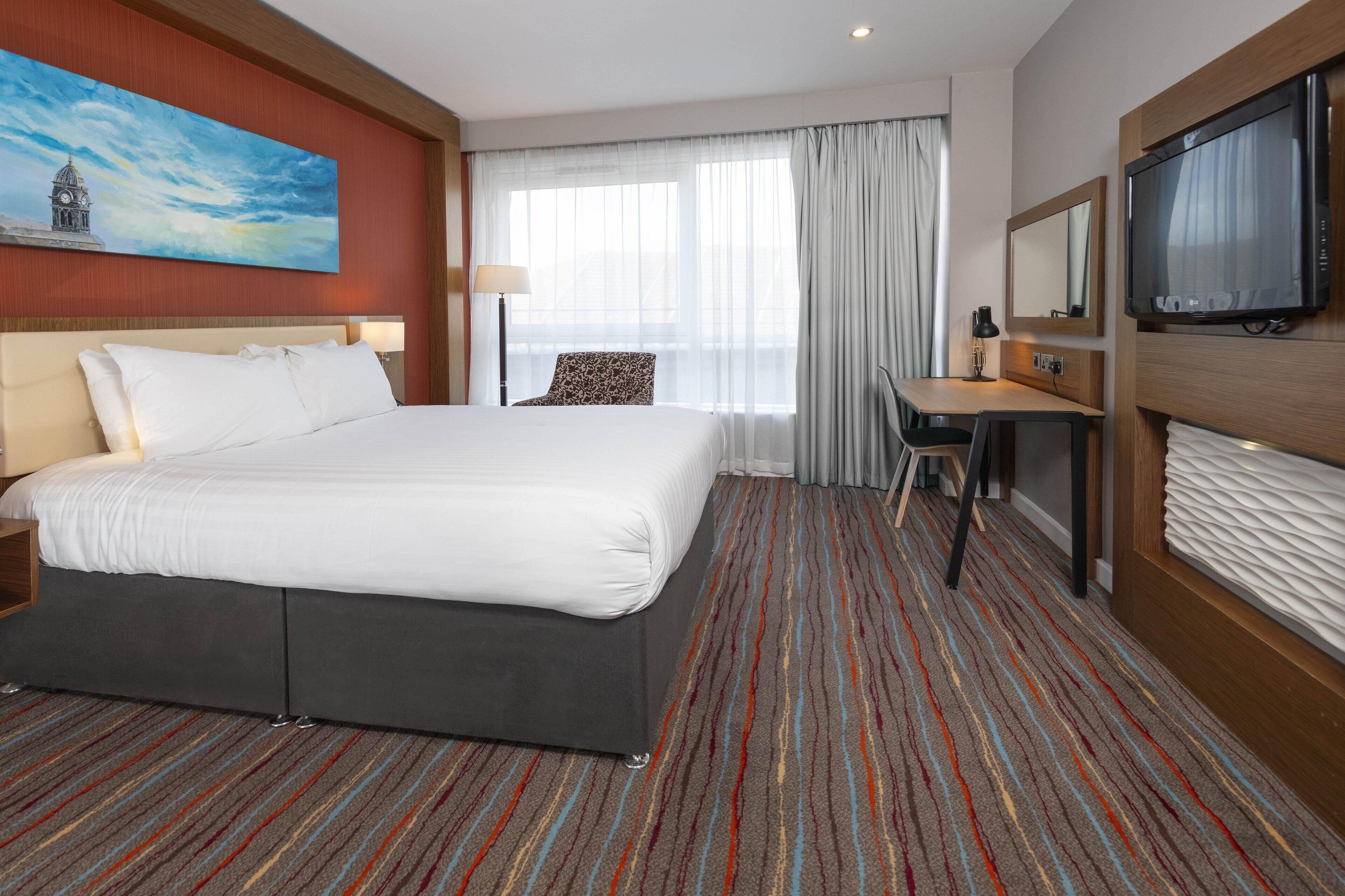 Une chambre d’hôtel avec un grand lit, un bureau, une télévision et un tableau représentant un bâtiment accroché au mur.
