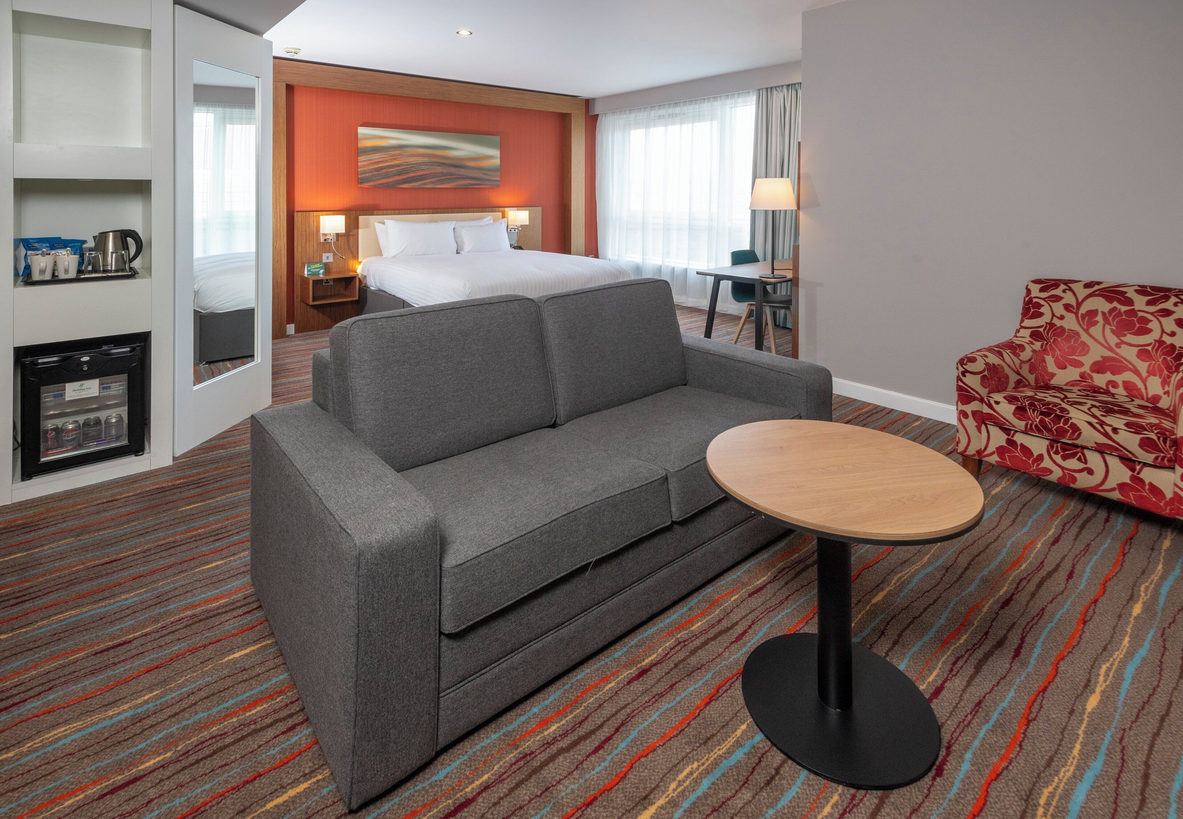 Habitación de hotel con sofá gris, una pequeña mesa redonda, un sillón estampado de flores, una cama con ropa blanca y una obra de arte colgada en la pared, además de un minibar.