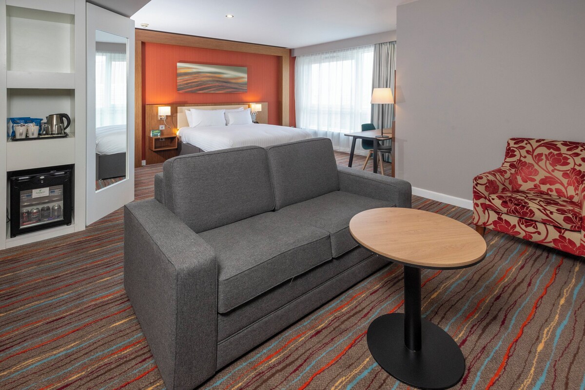 A hotel room with a grey sofa, a small round table, a floral armchair, a bed with white linens, a wall-mounted artwork, and a minibar.