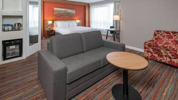 Habitación de hotel con sofá gris, una pequeña mesa redonda, un sillón estampado de flores, una cama con ropa blanca y una obra de arte colgada en la pared, además de un minibar.