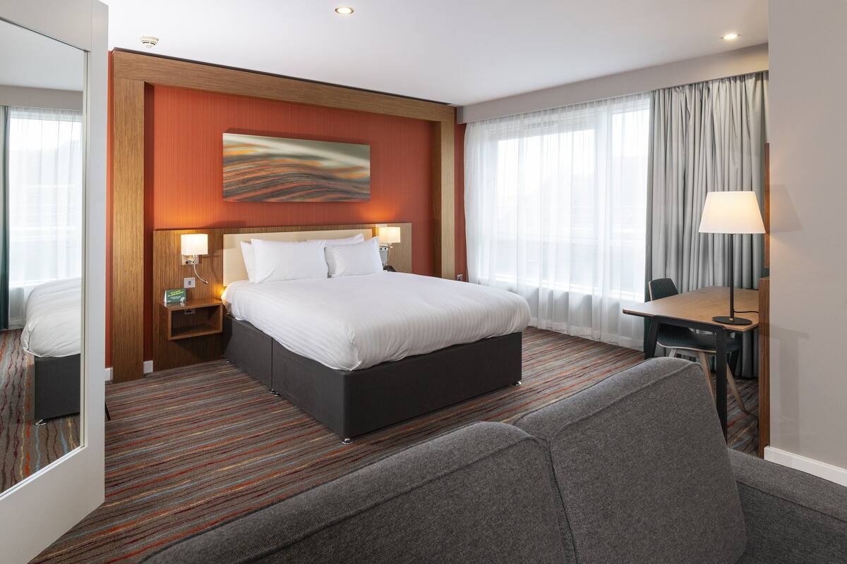 Una habitación de hotel con una cama grande, un escritorio, una silla, una lámpara y un cuadro en la pared.