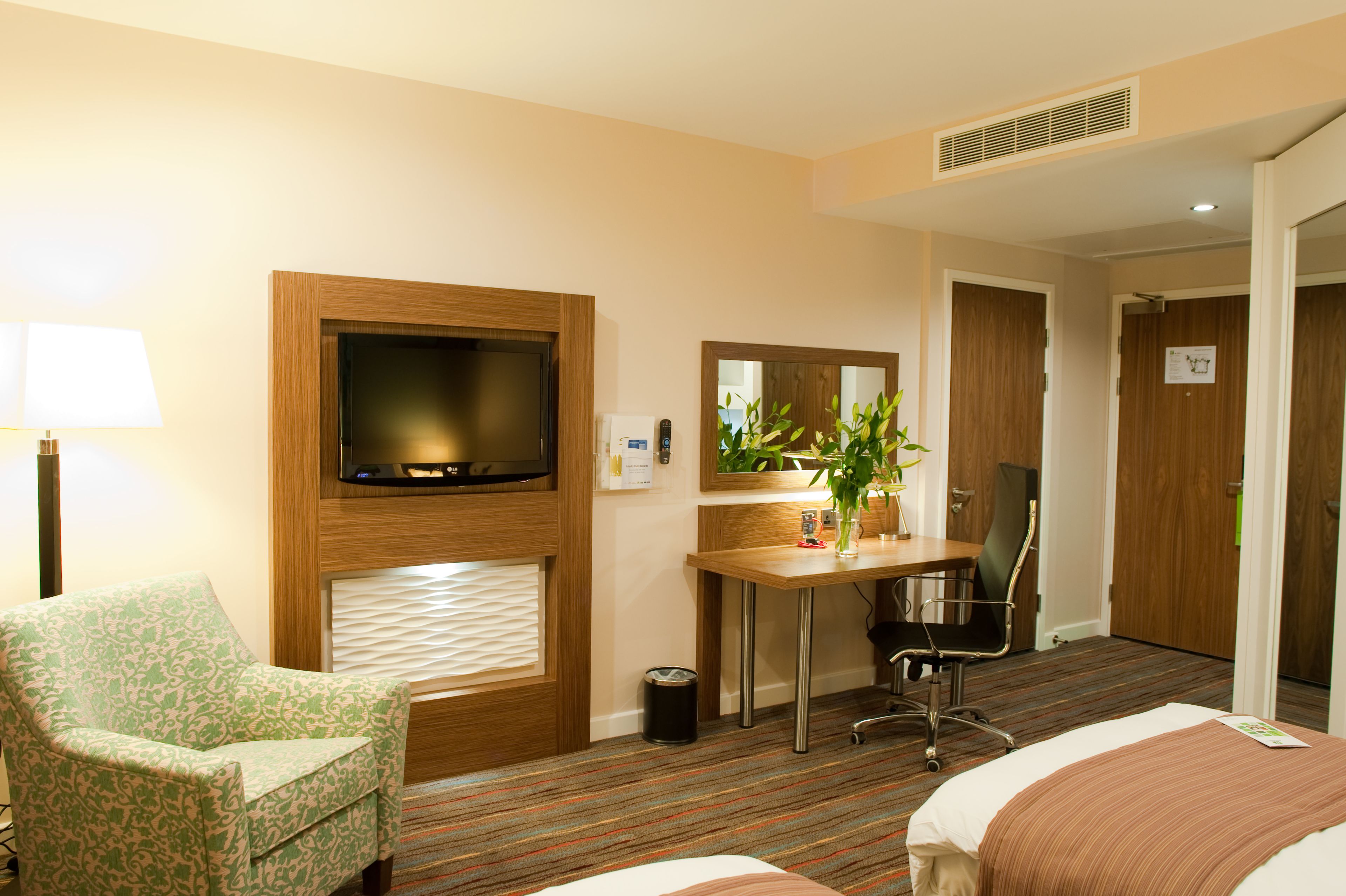 A hotel room with a bed, a desk with a chair, a television, a plant, and a mirror.