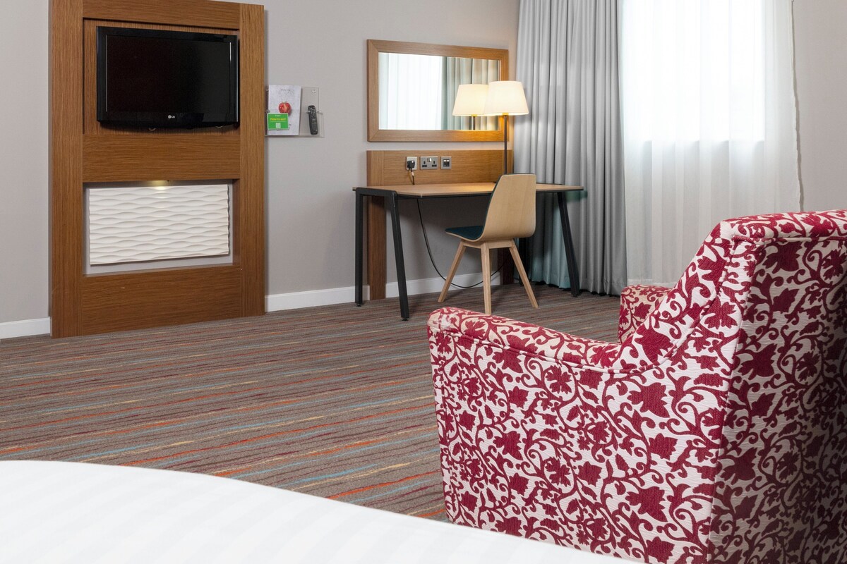 A hotel room with a bed, a desk, a chair, a television, a mirror, and a window with curtains.