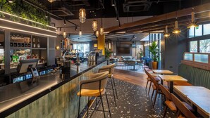 A modern bar area with a counter, bar stools, and pendant lighting. There are seating areas with tables and chairs, and a large window providing natural light. - Holiday Inn Derby Riverlights by IHG (Derby)