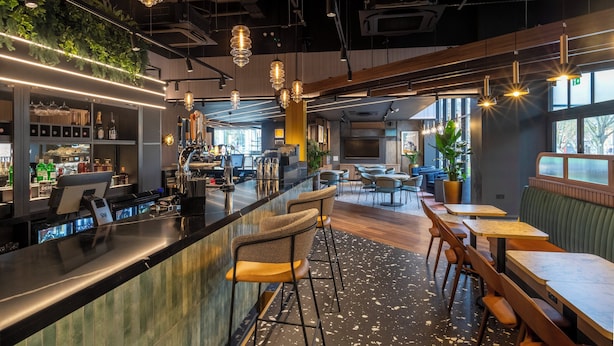 A modern bar area with a counter, bar stools, and pendant lighting. There are seating areas with tables and chairs, and a large window providing natural light.