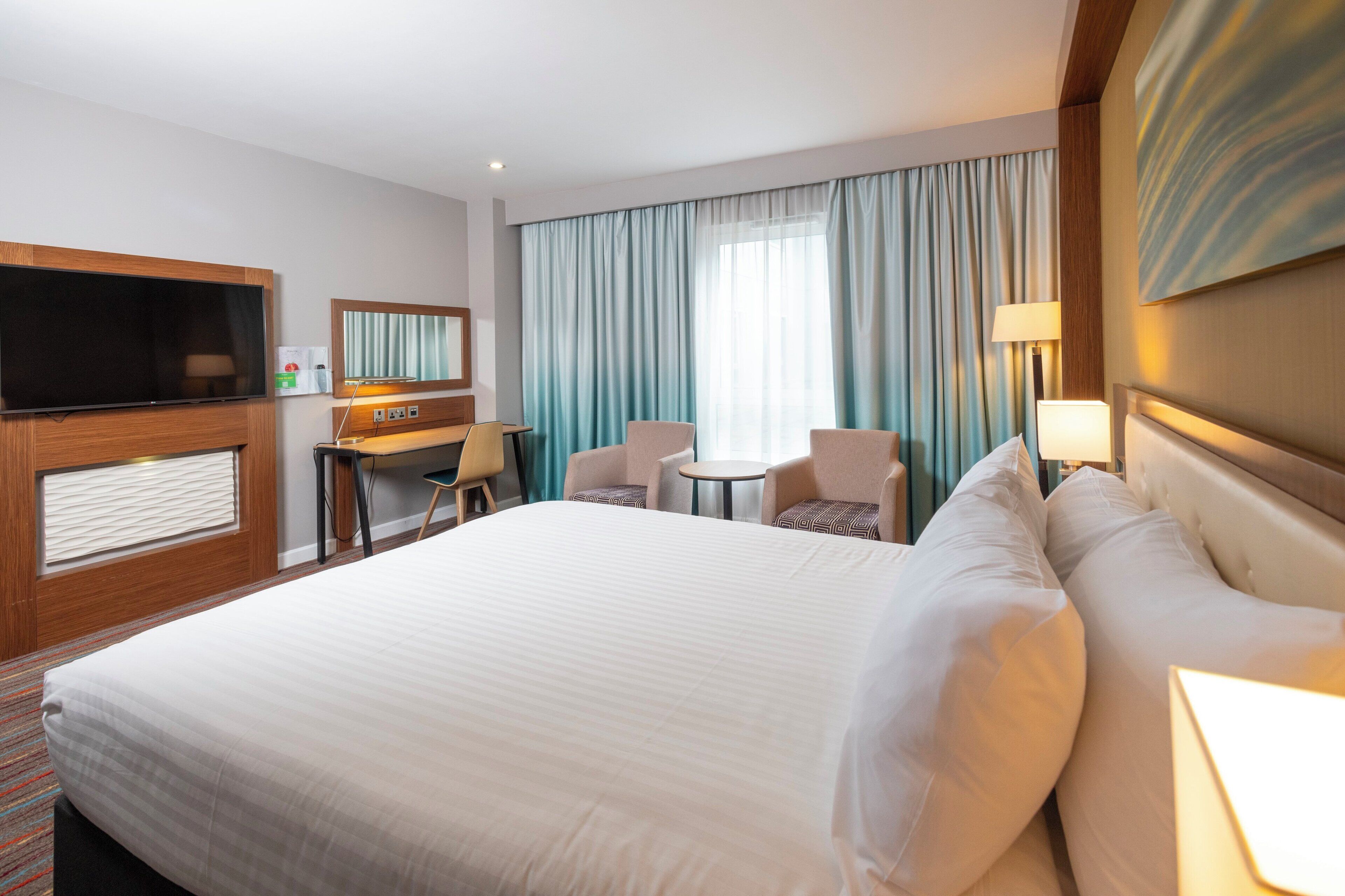 A hotel room with a large bed, a desk with a chair, a television, and a window with curtains.