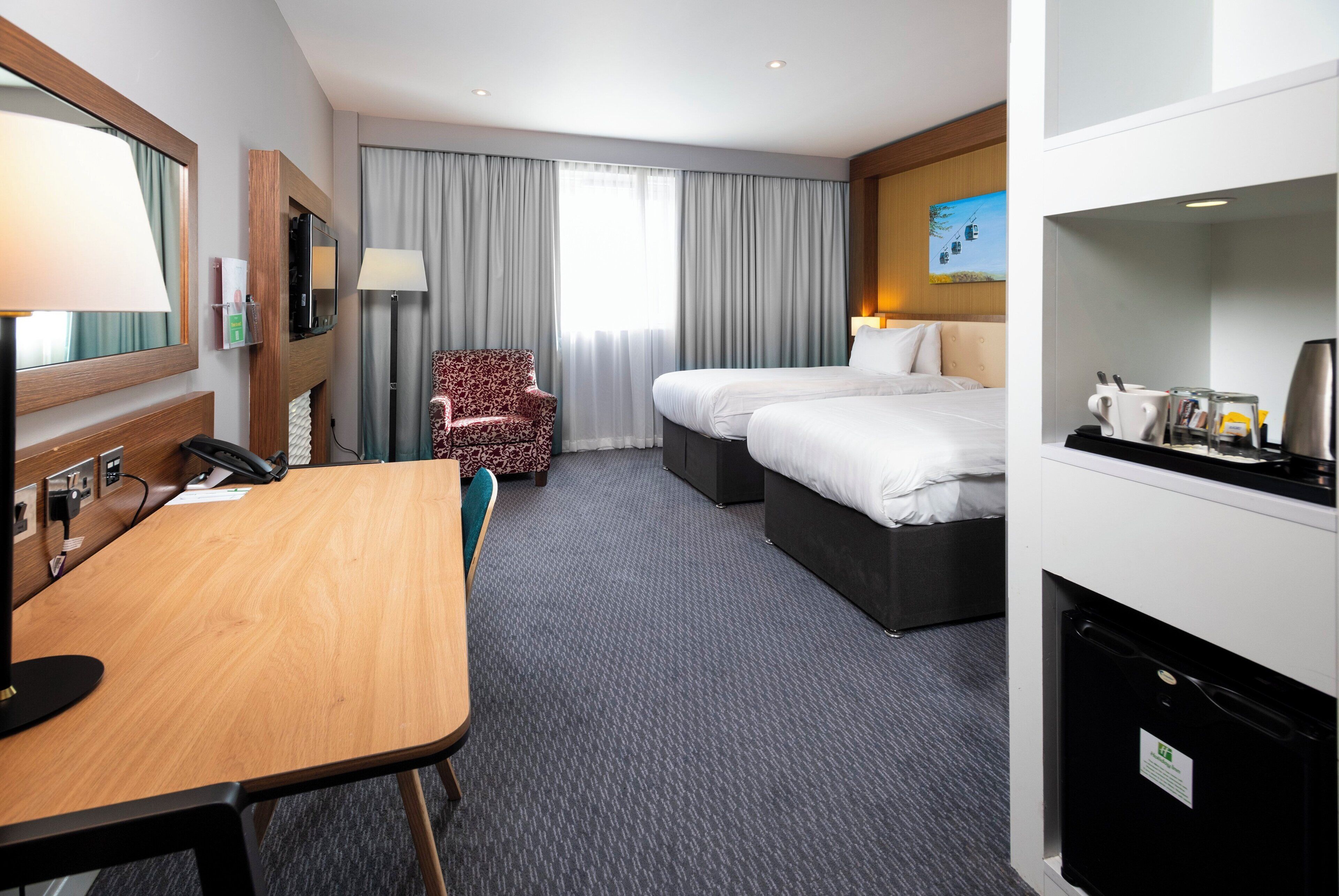A hotel room with a desk, two beds, a chair, a TV, and a minibar.