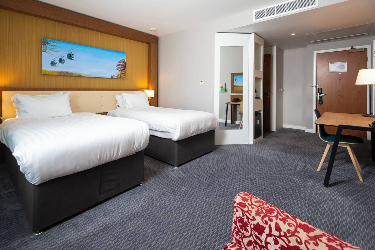 Habitación de hotel con dos camas, un escritorio, una silla y un ventanal con vistas a una montaña y un teleférico.