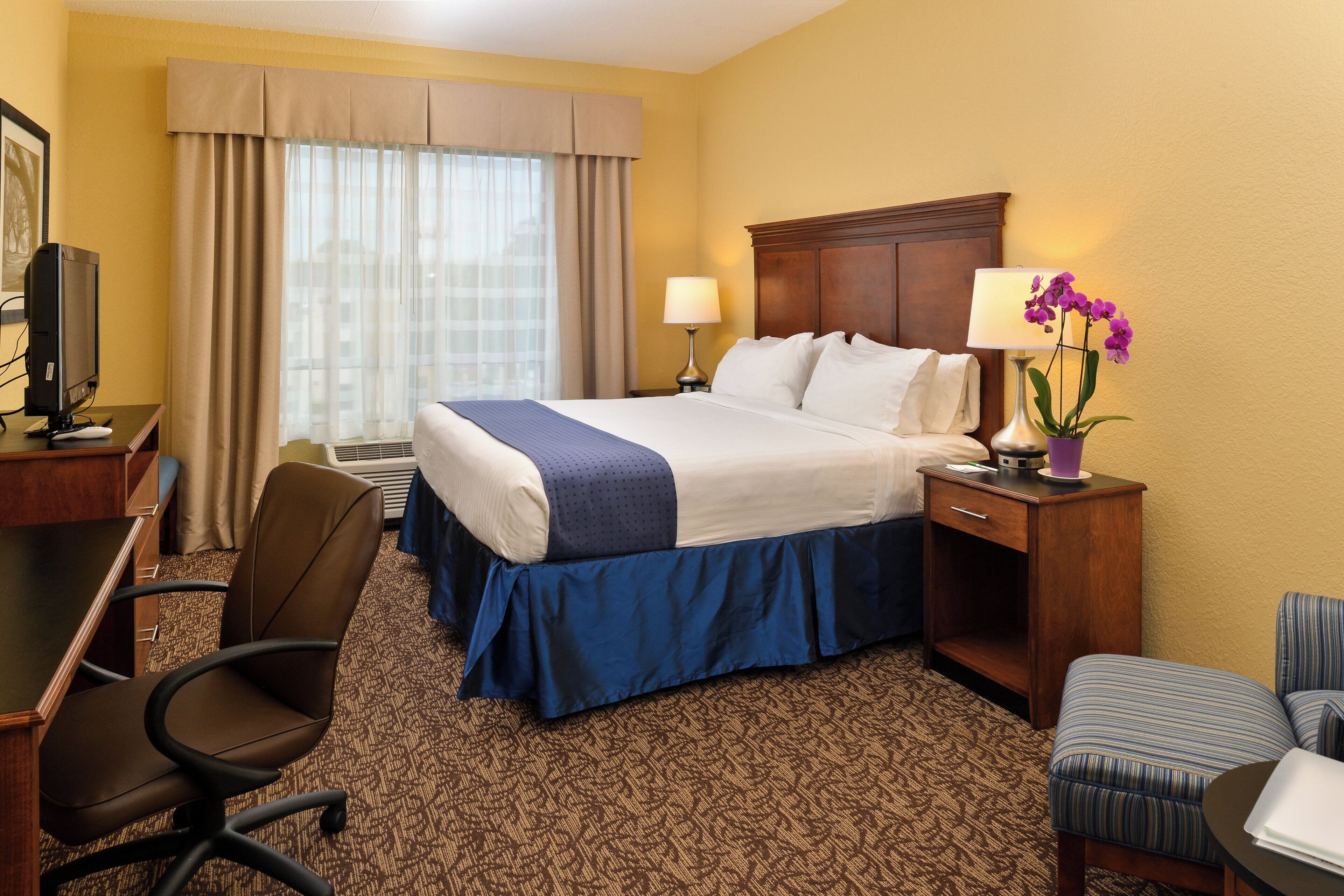 A hotel room with a bed, desk, chair, lamp, and an orchid plant.