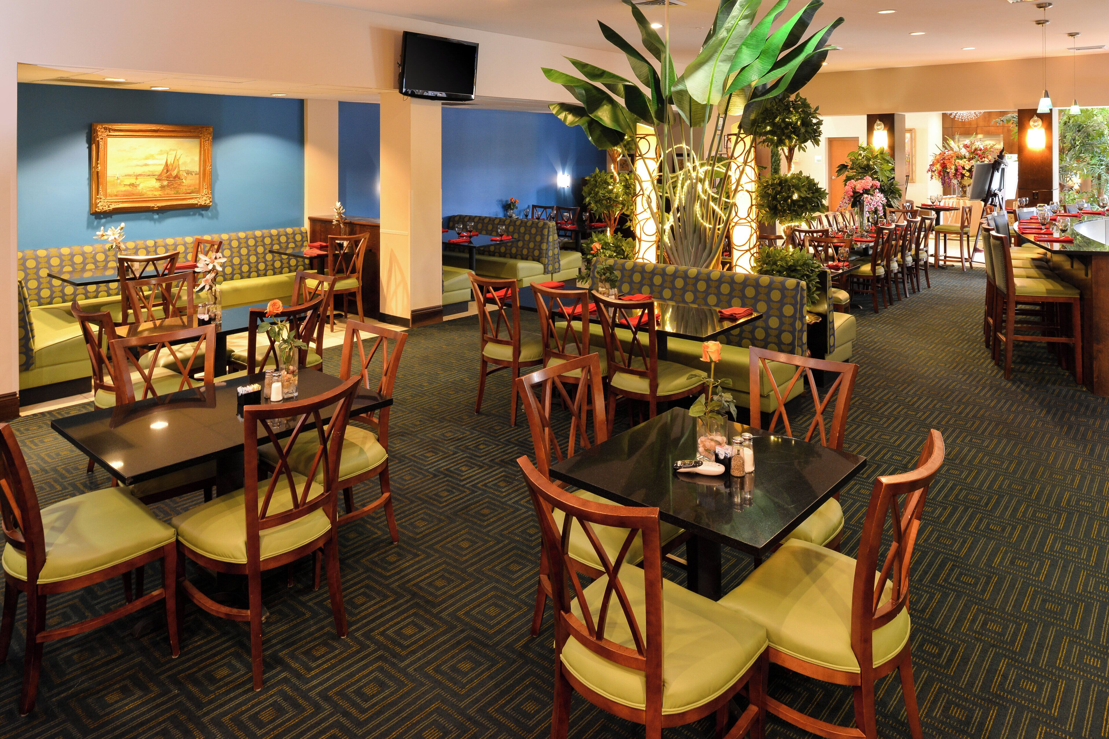 A hotel dining area with multiple seating options including booths and tables, a television mounted on the wall, and various plants adding greenery.